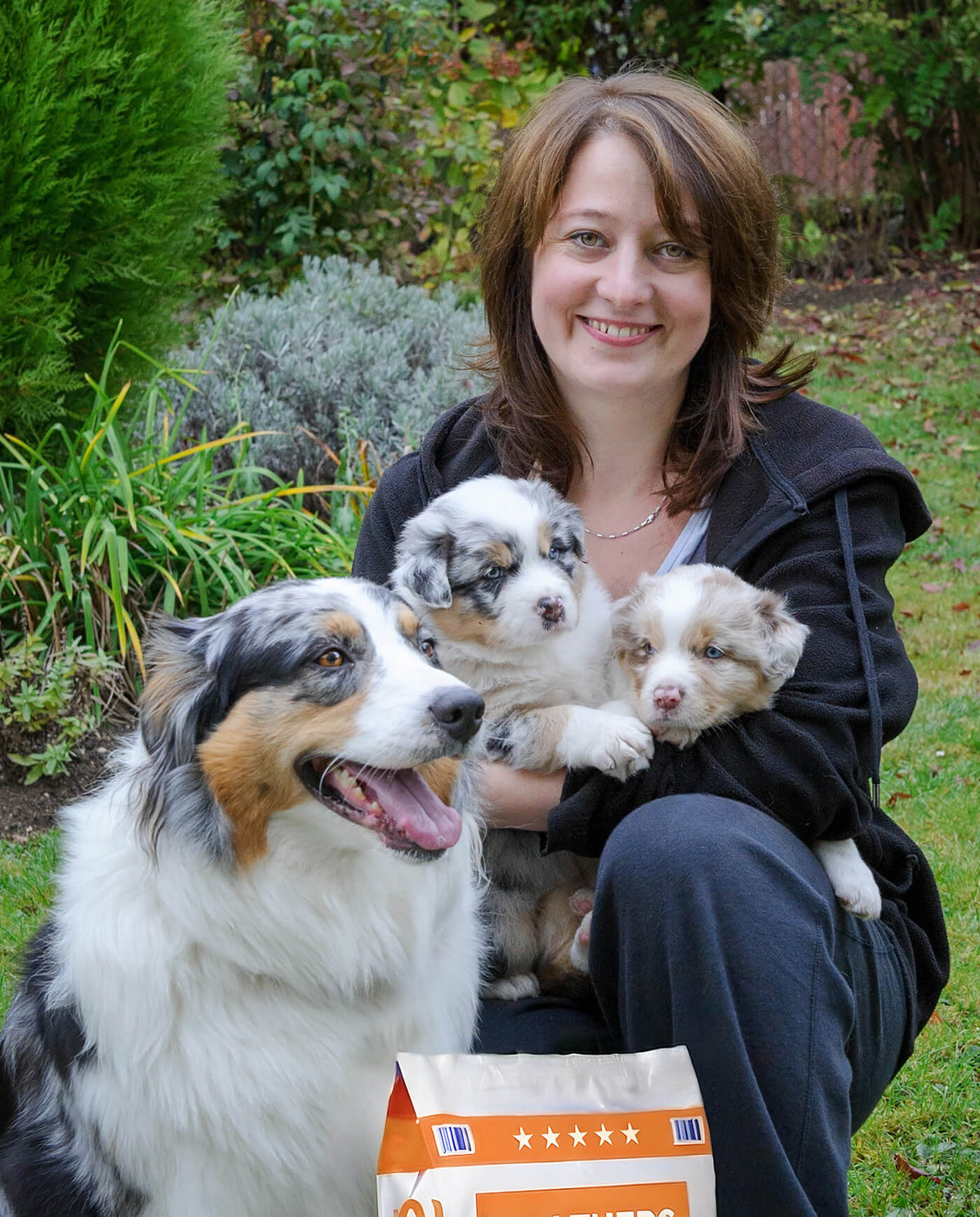 Woman holding three puppies with a dog in a garden setting